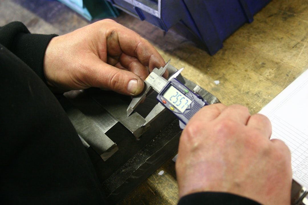 A machinist's hands measuring a metal part with a digital caliper at a workbench